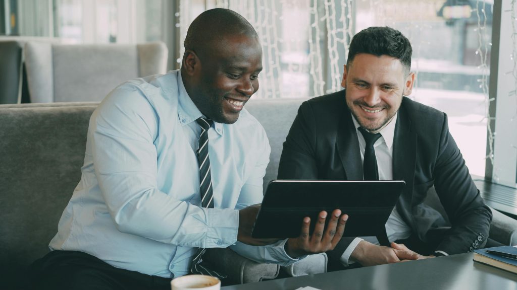 Home Two businessmen happily collaborating on a digital tablet in a modern cafe setting.